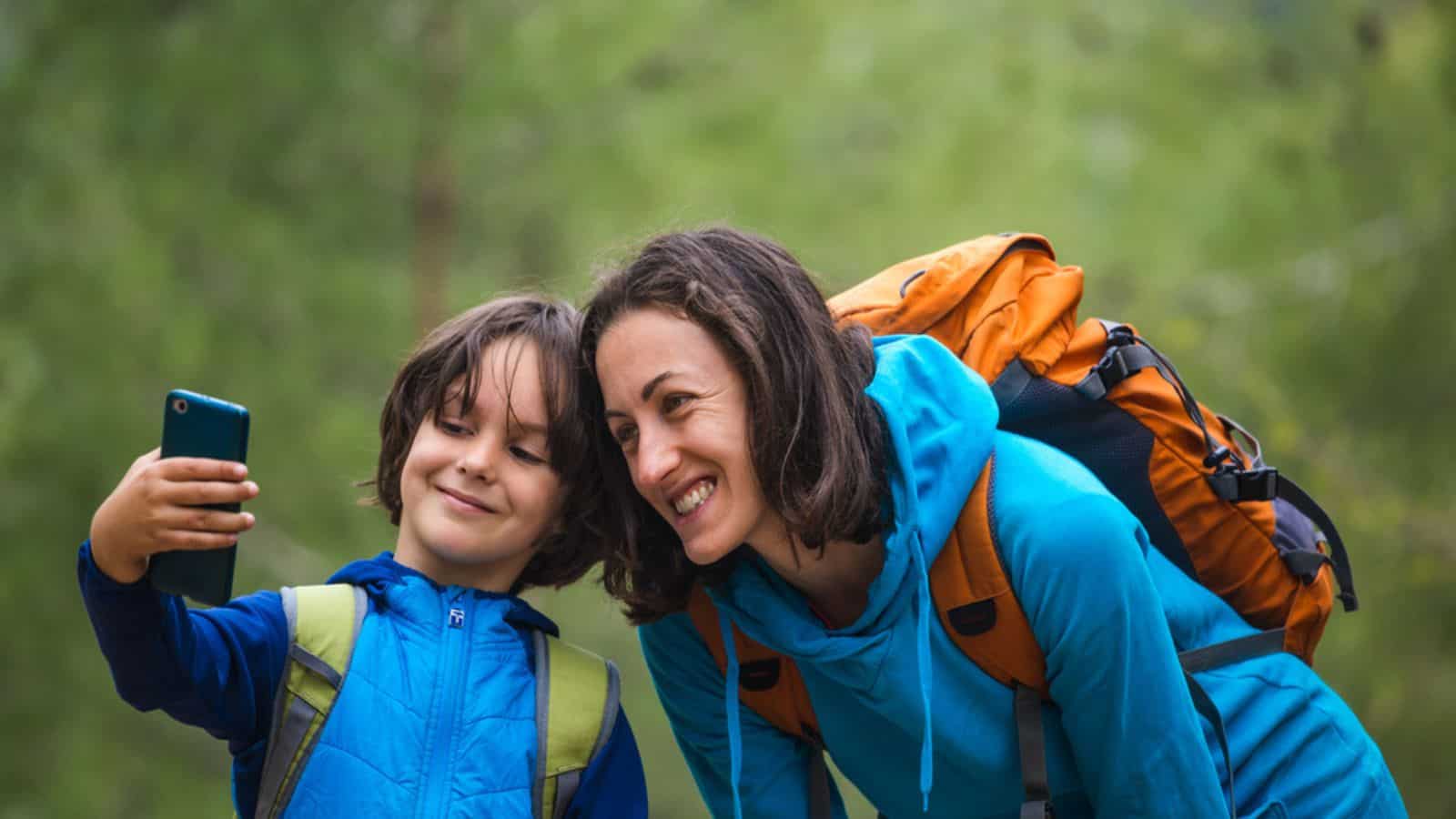 A child with a backpack takes a selfie on a smartphone with mom on the background of a mountain forest, A boy travels with mother, A woman is photographed with her son, Hiking with children