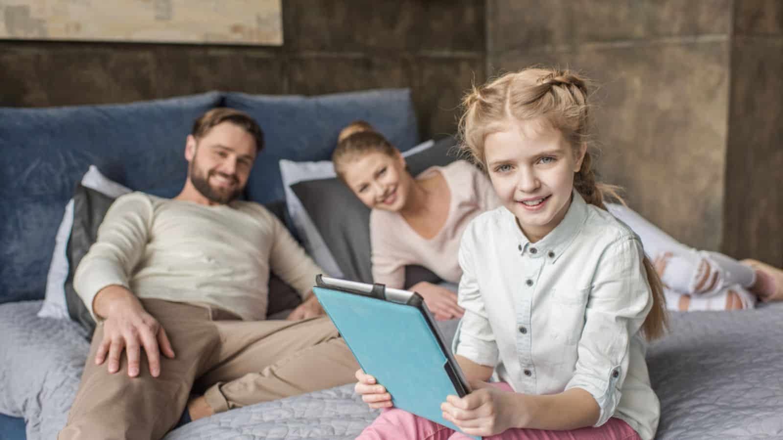 Adorable daughter using digital tablet and lying on bed with parents