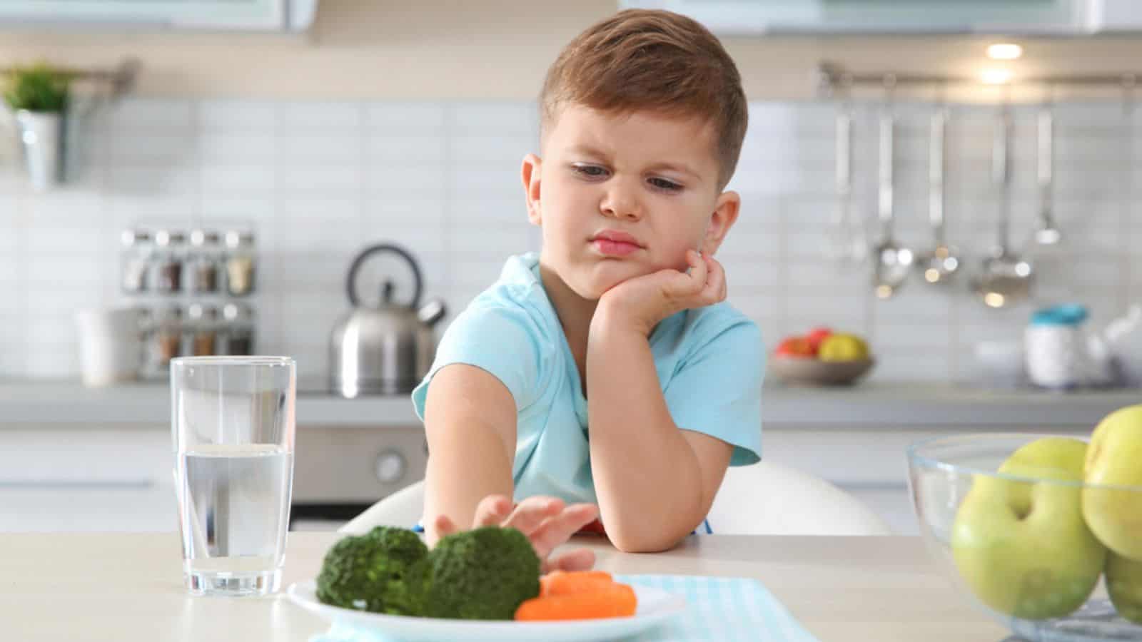 Adorable little boy refusing to eat vegetables at table in kitchen