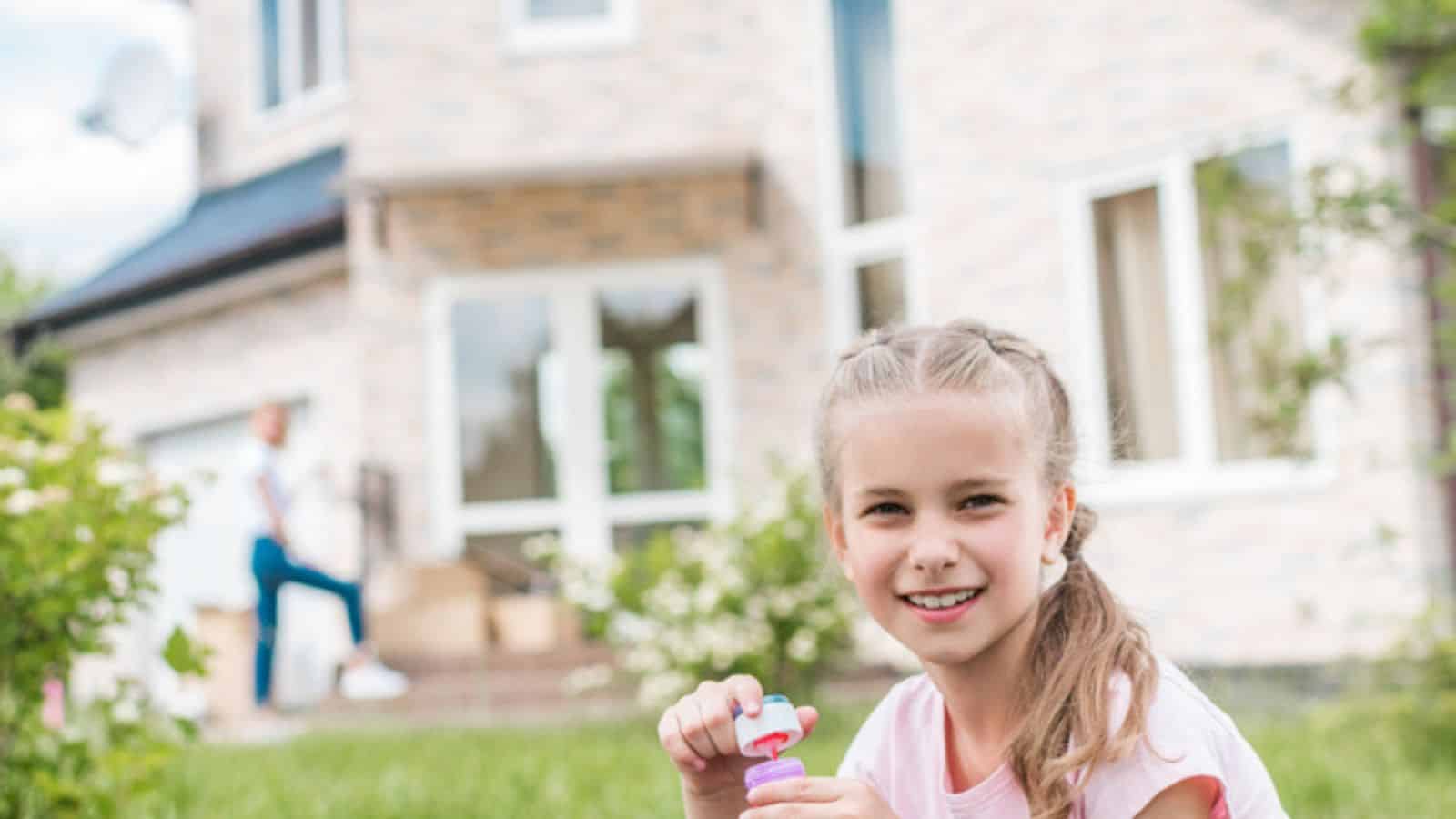 Adorable little child with soap bubbles sitting on lawn while her mother standing behind in front of house