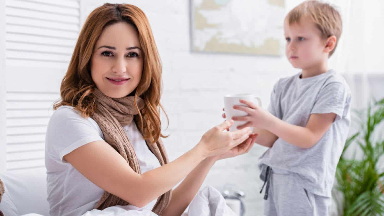 Adorable son giving cup of tea to sick mommy in bedroom