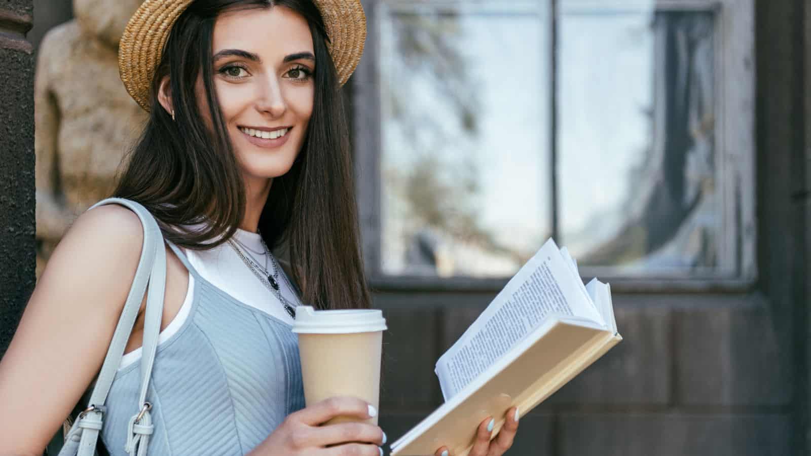 Attractive girl with paper cup reading book and smiling at camera