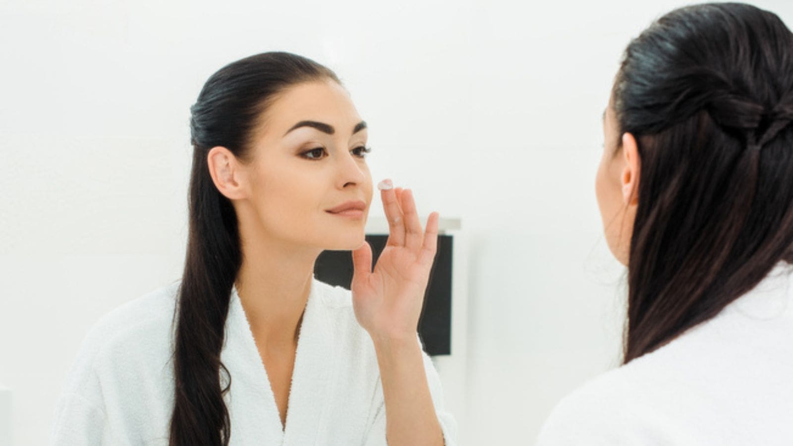 Beautiful woman in bathrobe looking at mirror and applying face cream