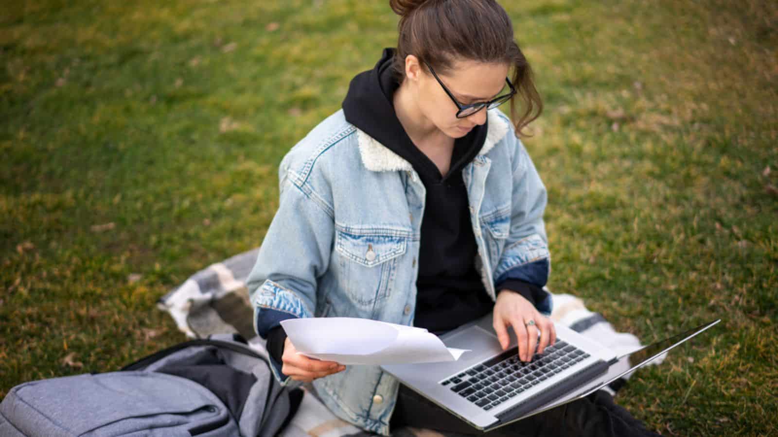 Beautiful young female student sitting on the grass using laptop in the park