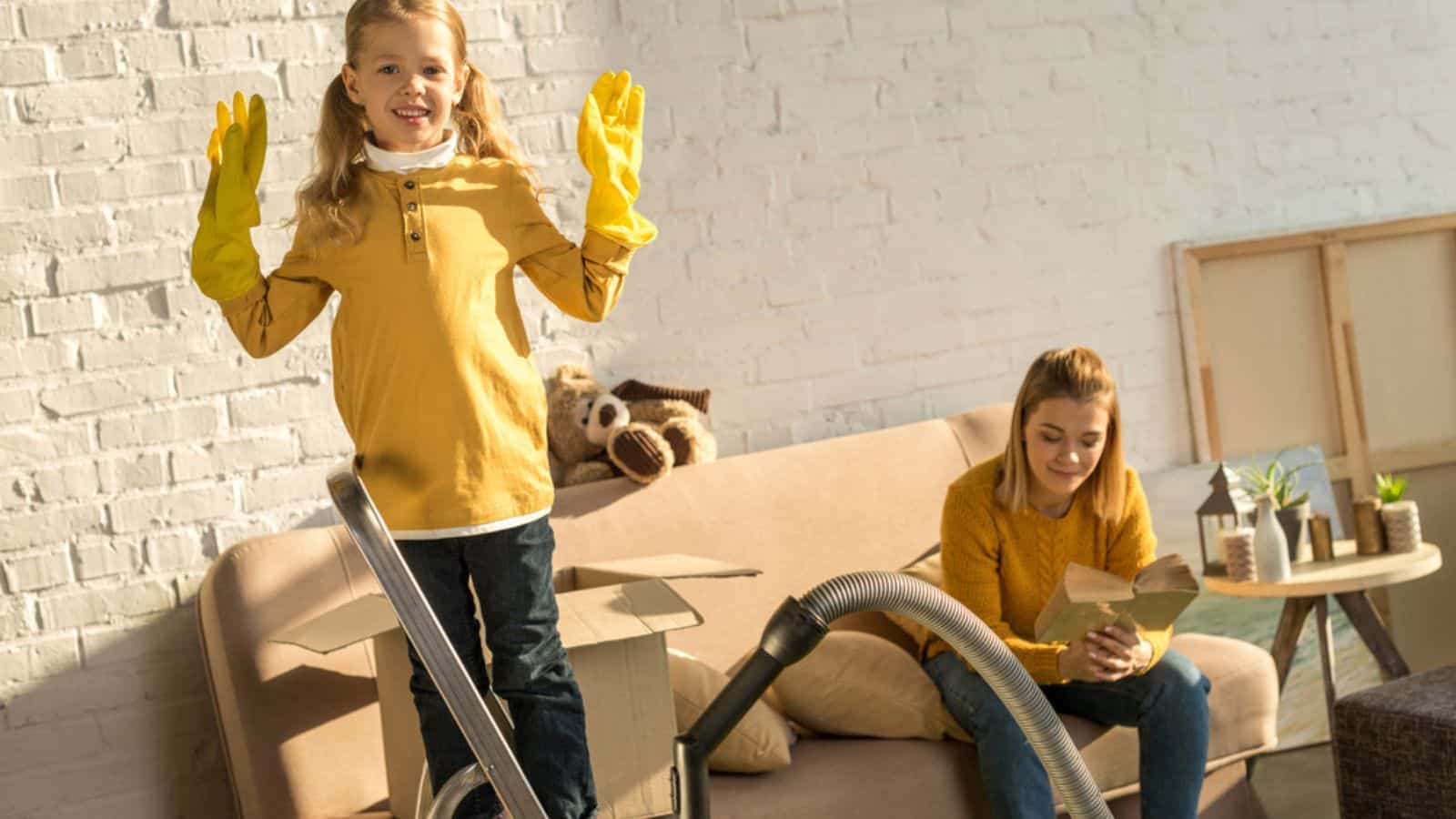 Child in rubber gloves smiling at camera while mother reading book on soda cleaning the house with vacuum