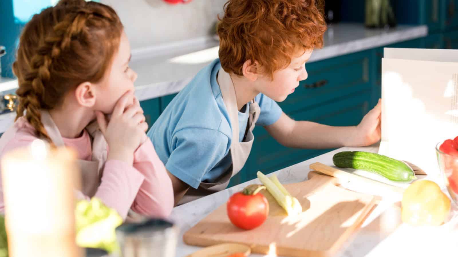 Children in aprons reading coobook while preparing vegetable
