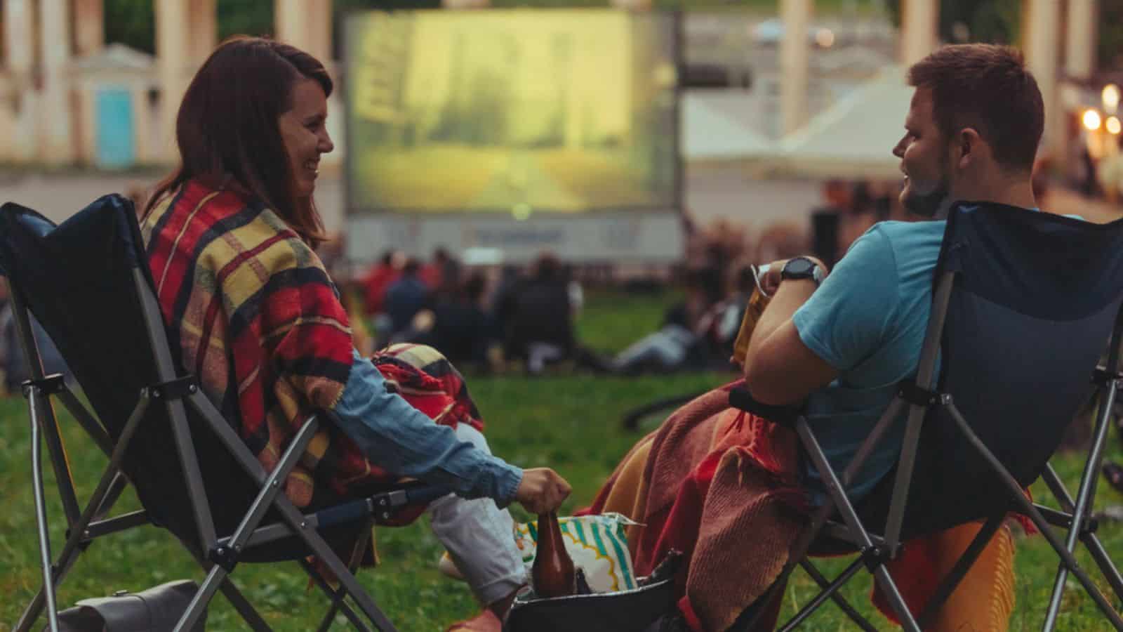 Couple sitting in camp-chairs in city park looking movie outdoors at open air cinema