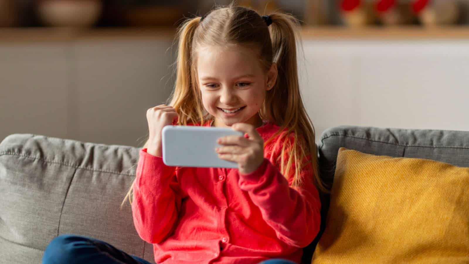 Cute Little Girl Playing Games On Smartphone At Home And Celebrating Success