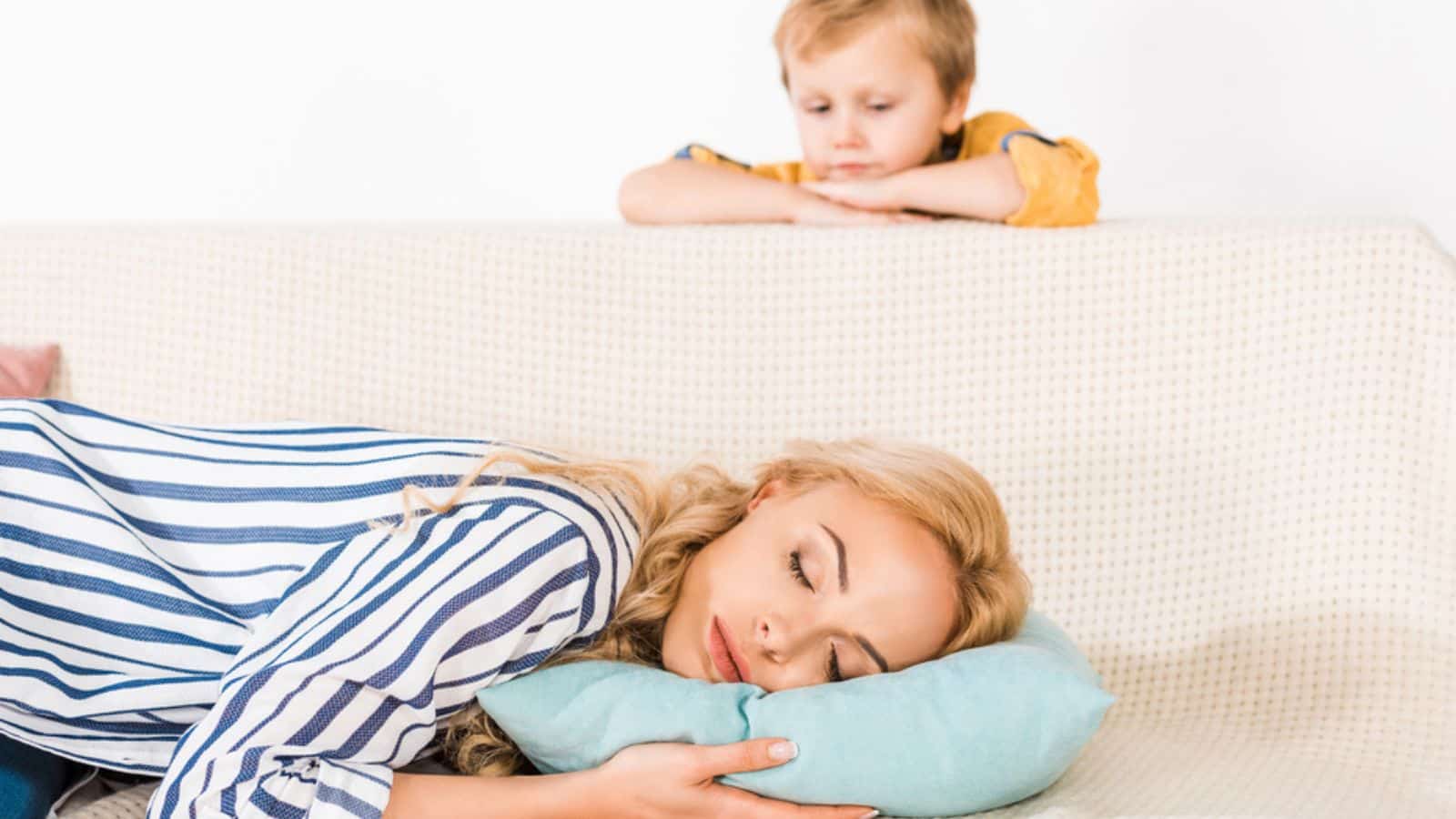 Cute little boy looking at mother sleeping on sofa at home