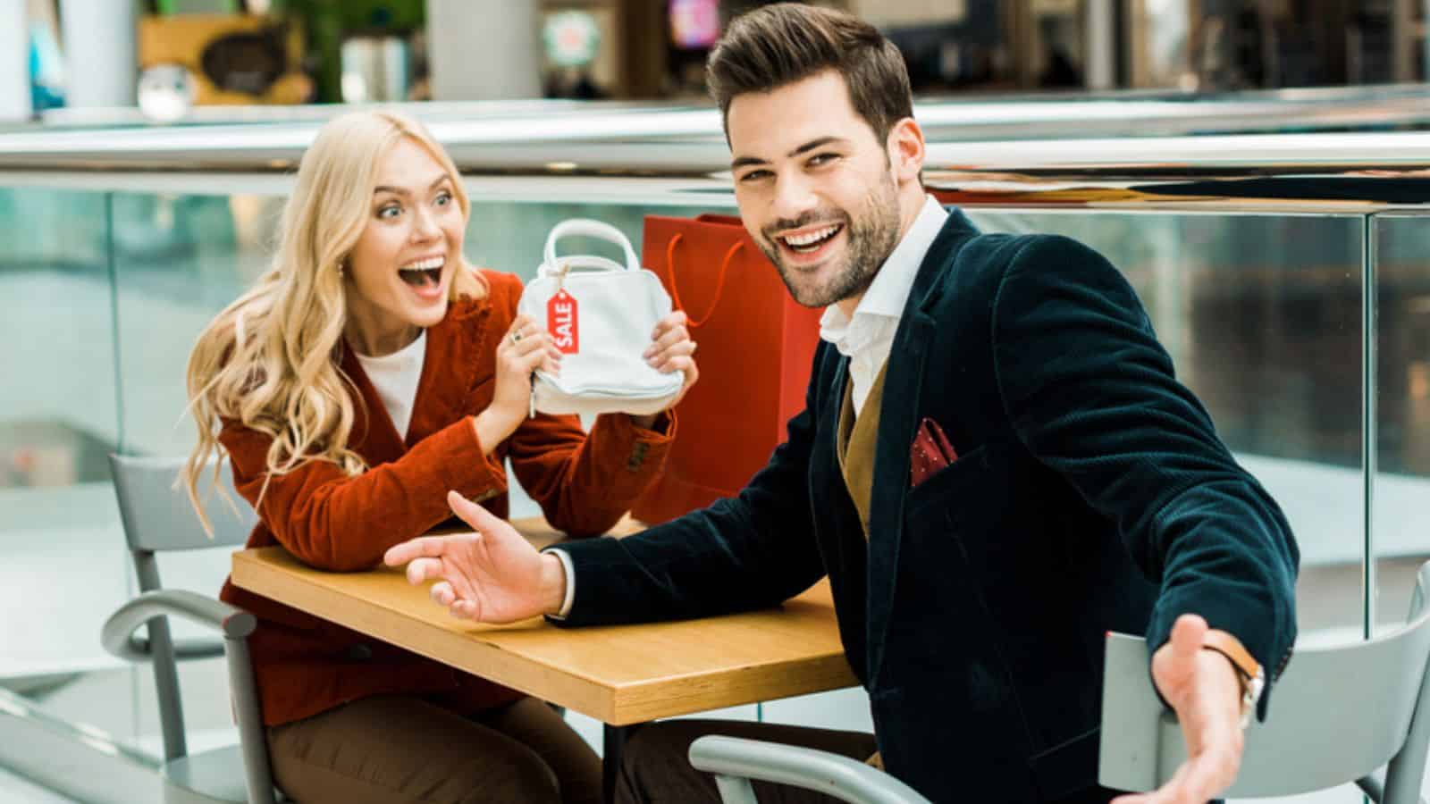Excited female shopaholic showing bag with sale tag to laughing boyfriend