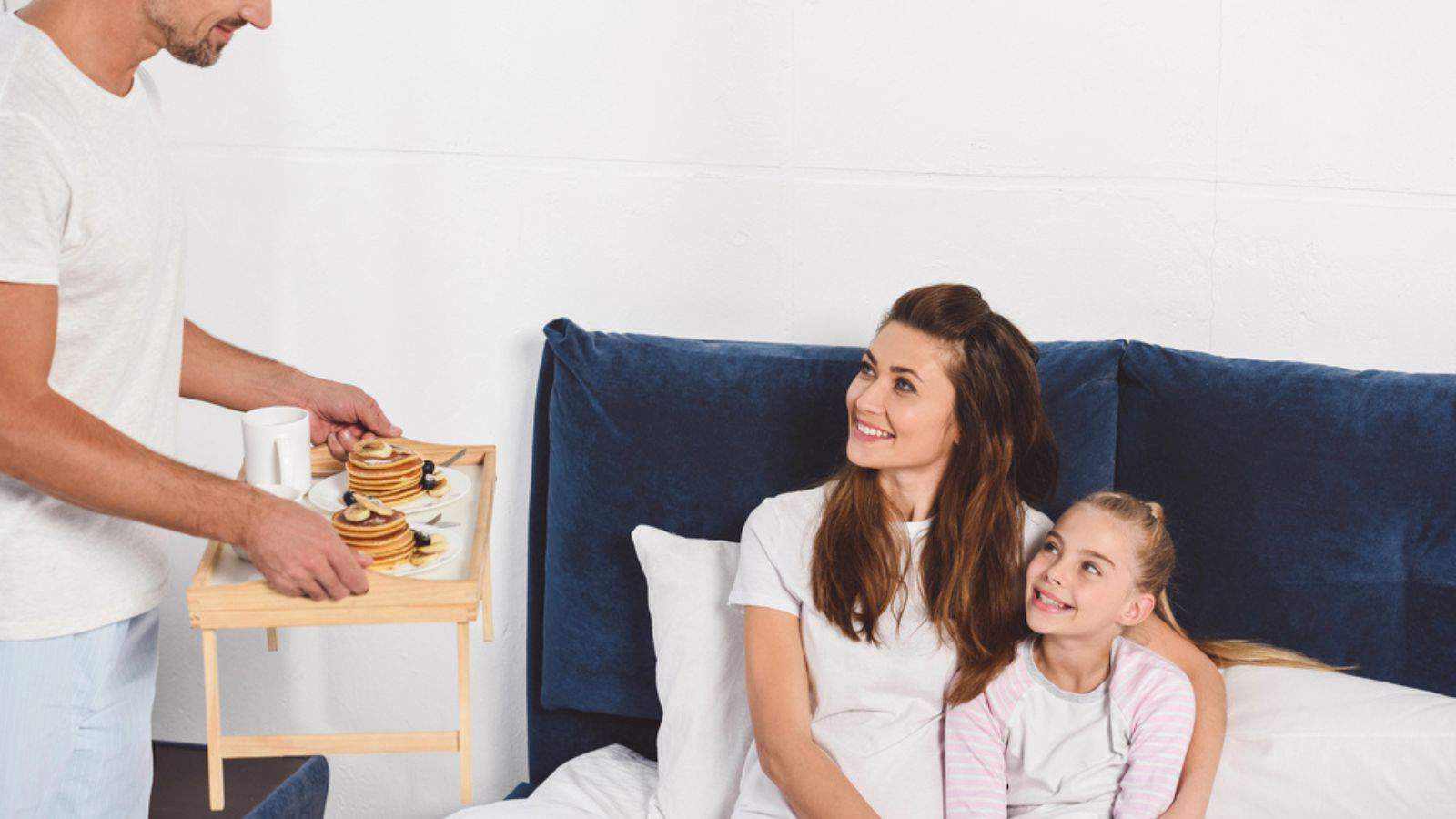 Father holding wooden tray with pancakes while daughter and wife smiling and sitting in bed
