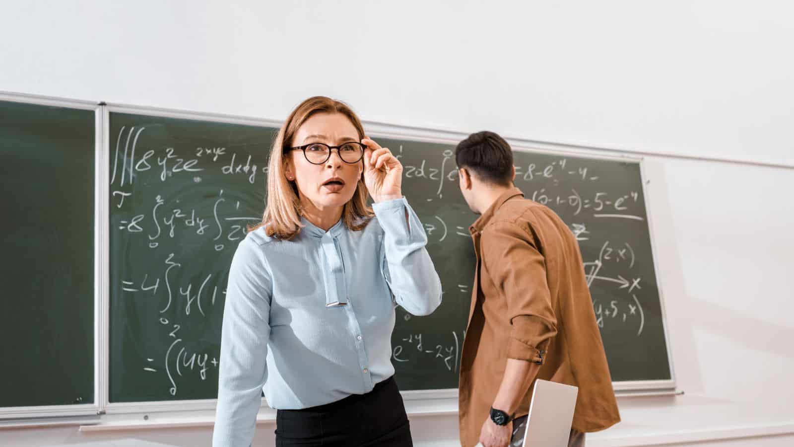 Female teacher standing near desk and looking surprised near student