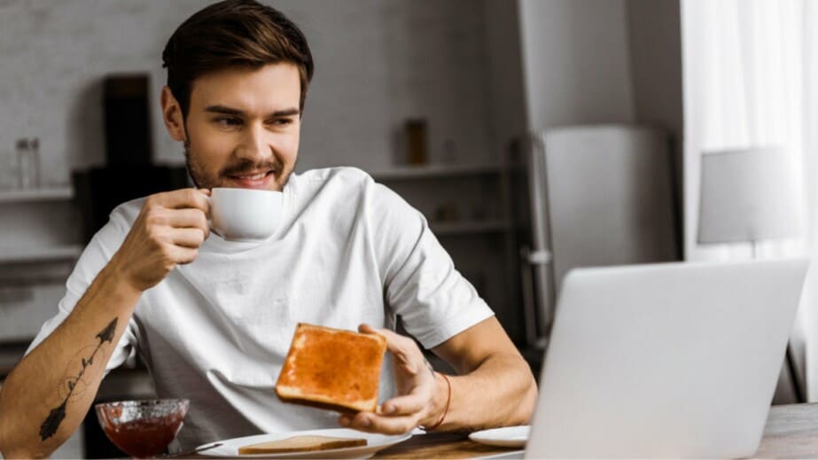 Grimacing young freelancer eating toast with jam and looking at laptop