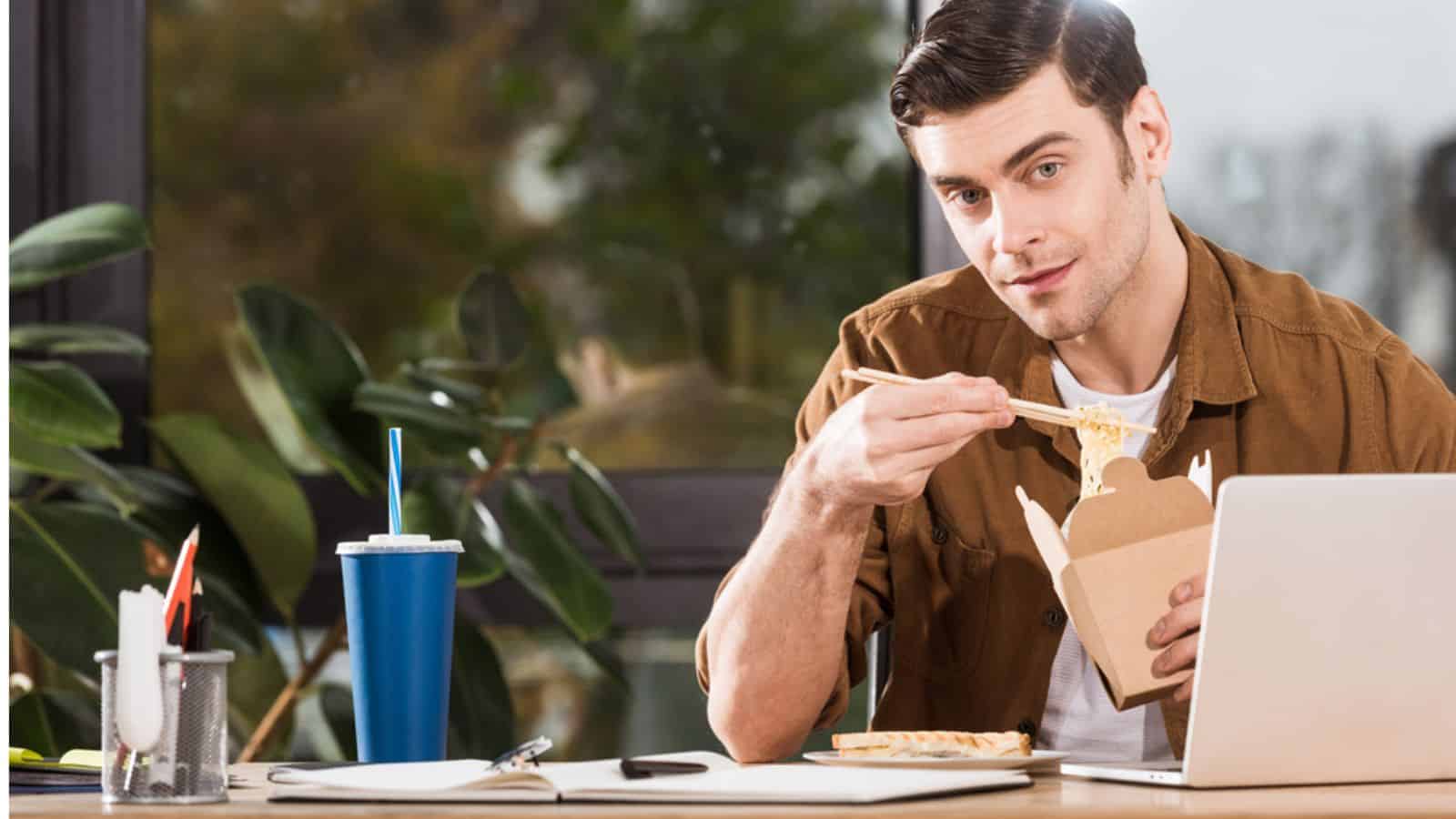 Handsome businessman eating take away noodles from box at office and looking at camera