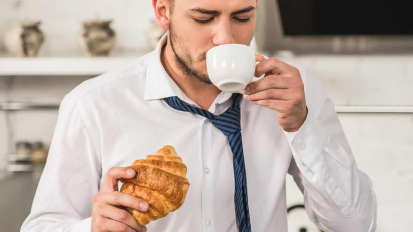 Handsome man drinking coffee and eating croissant in morning at kitchen