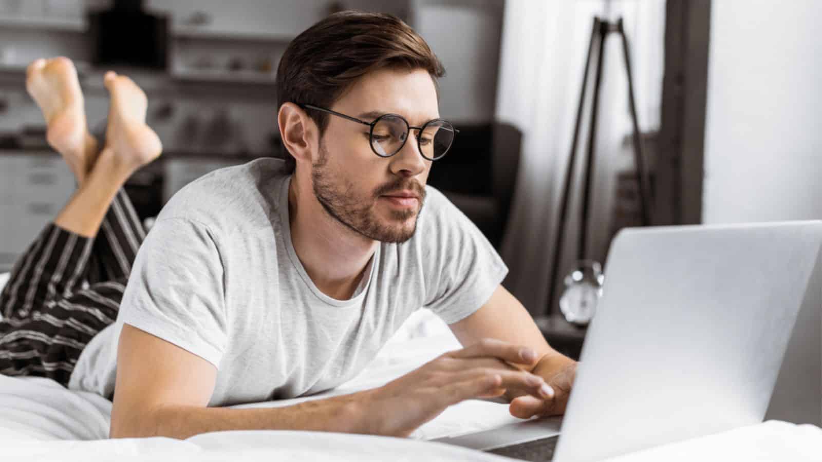Handsome young man in eyeglasses and pajamas using laptop on bed