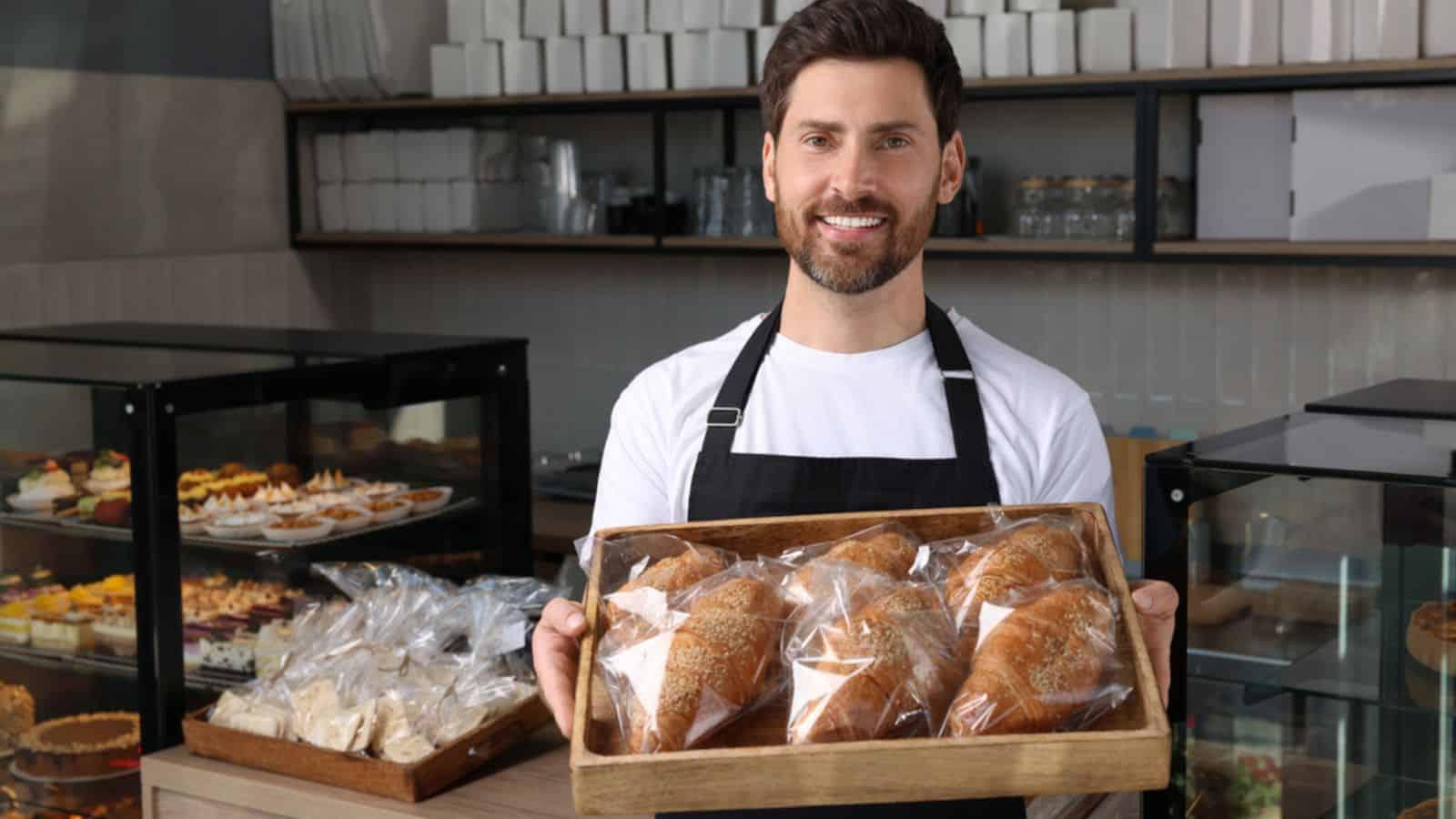 Happy man seller business owner holding tray with tasty croissants in bakery shop