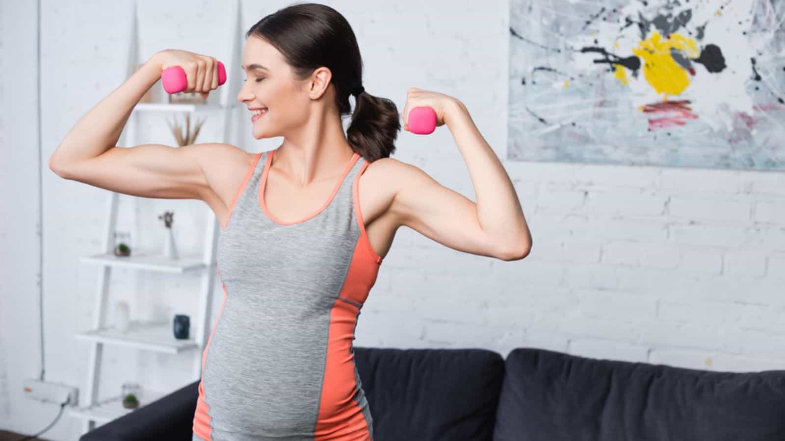 Happy pregnant woman exercising with pink dumbbells at home