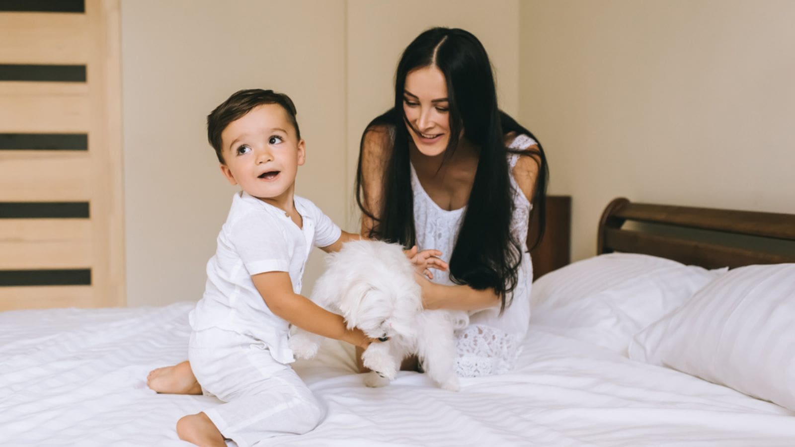 Happy young mother and child playing with bichon dog while sitting on bed at home