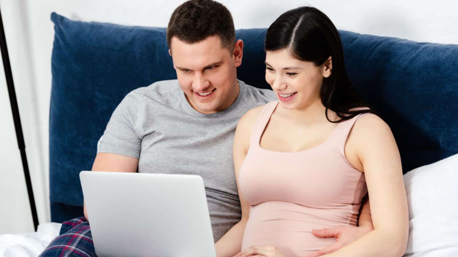 Happy young pregnant couple using laptop together on bed