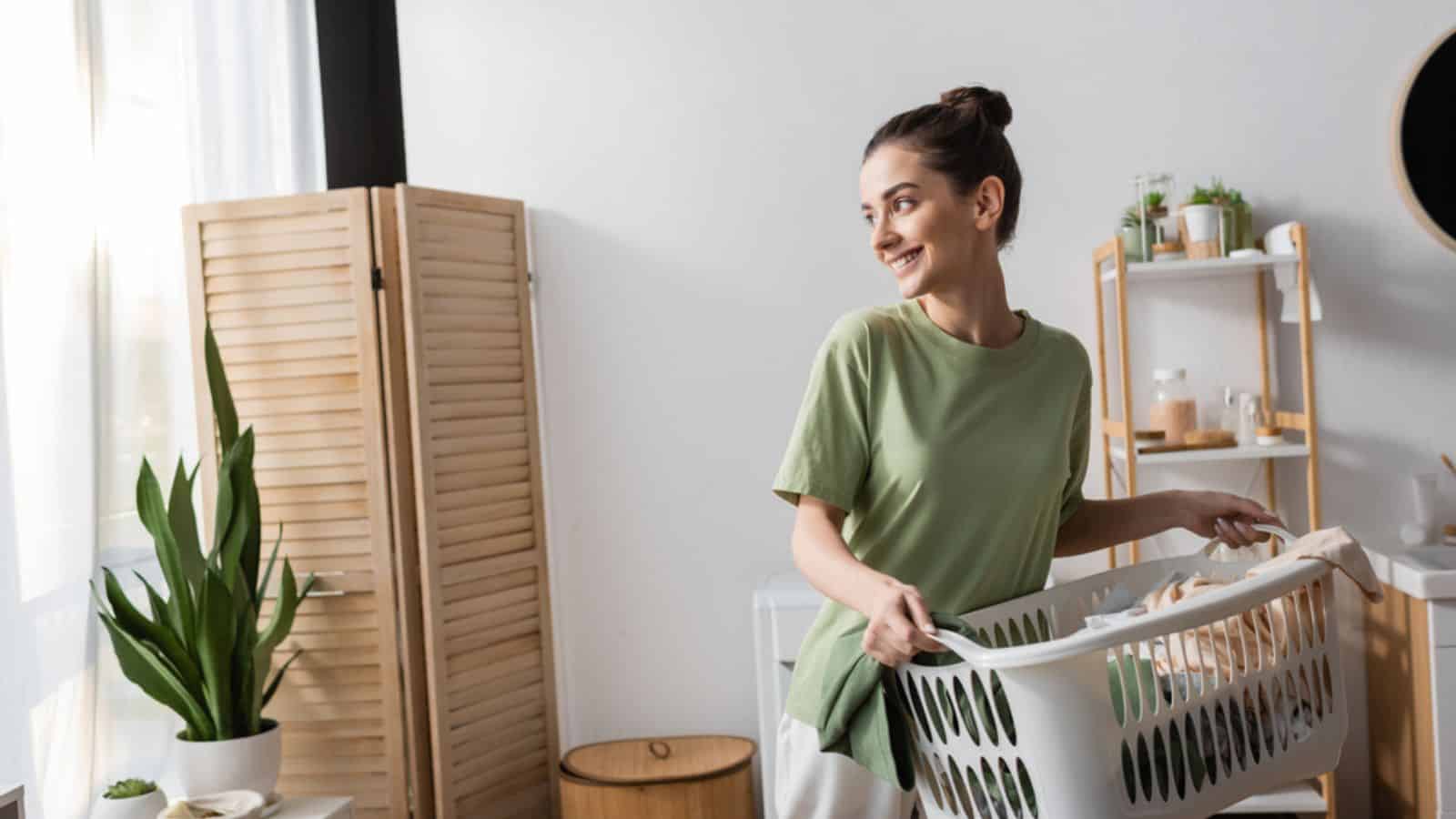 Happy young woman holding basket with clothes in laundry room