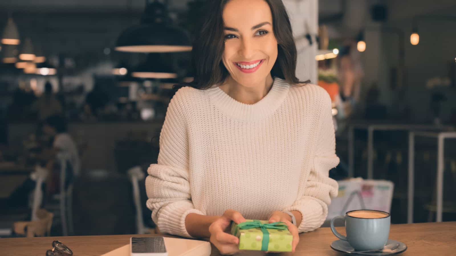 Happy young woman holding gift box at table with coffee cup