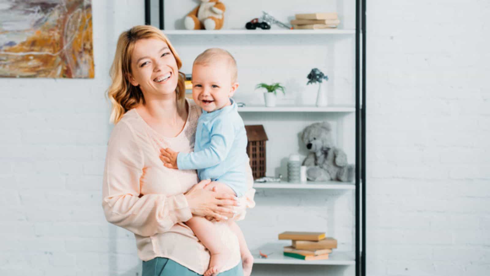 Laughing mother carrying her little child and looking at camera at bedroom