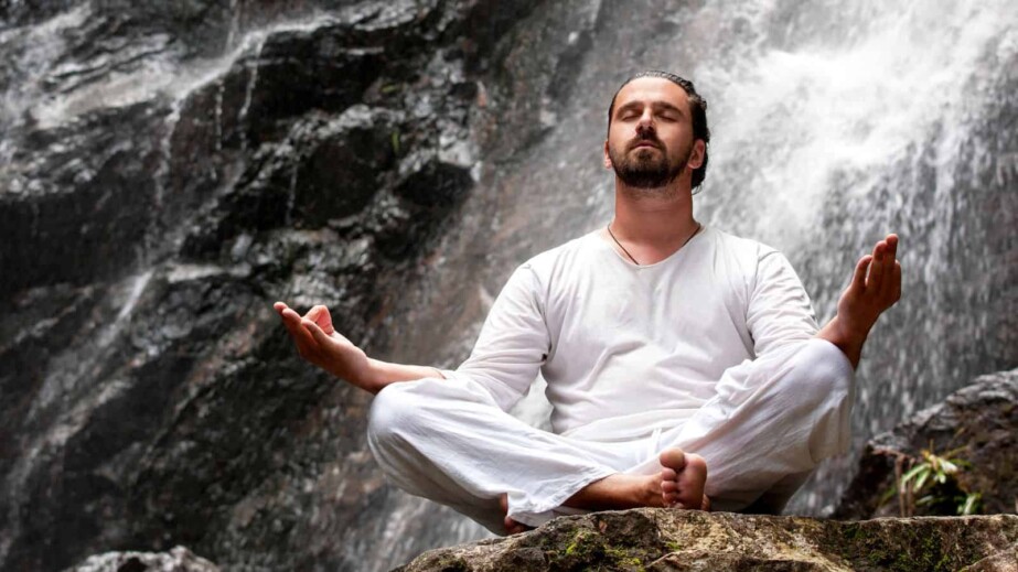 Man Sitting in Meditation Yoga on Rock at Waterfall