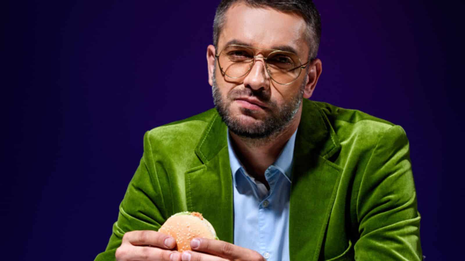 Man in velvet jacket eating burger at table with french cries
