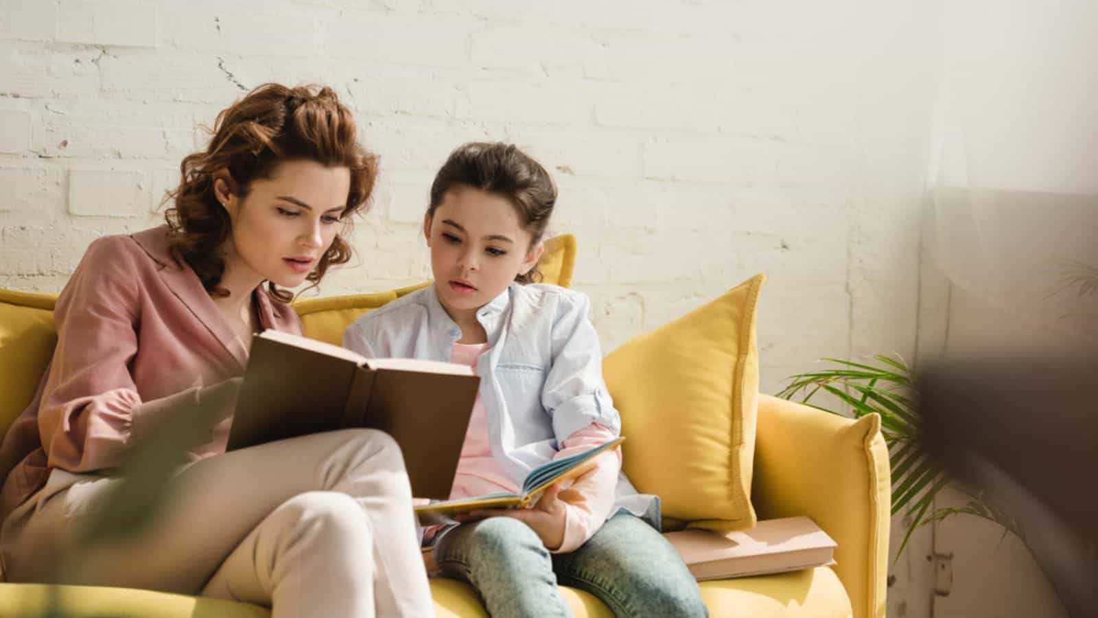 Mother and daughter sitting on yellow sofa at home and reading