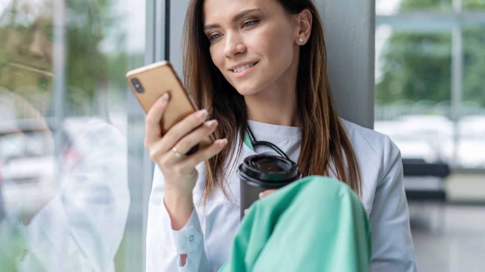Portrait of young woman doctor in white coat sitting while using smartphone