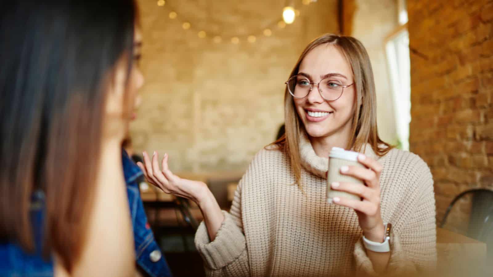 Positive woman in casual sweater with toothy smile and eyeglasses sitting in cafe