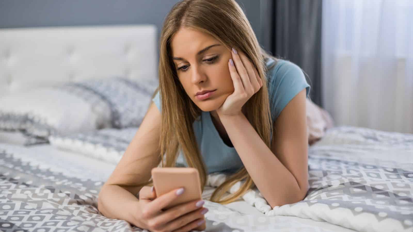 Sad woman holding phone while lying down on bed in her bedroom