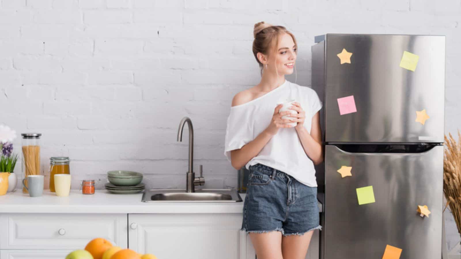 Selective focus of young blonde woman in white t-shirt and shorts holding cup while standing in kitchen