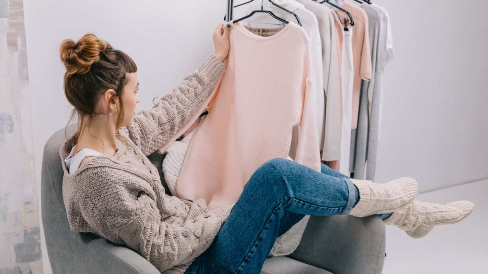 Side view of girl sitting on armchair and looking at shirt in hand thinking to declutter clothes