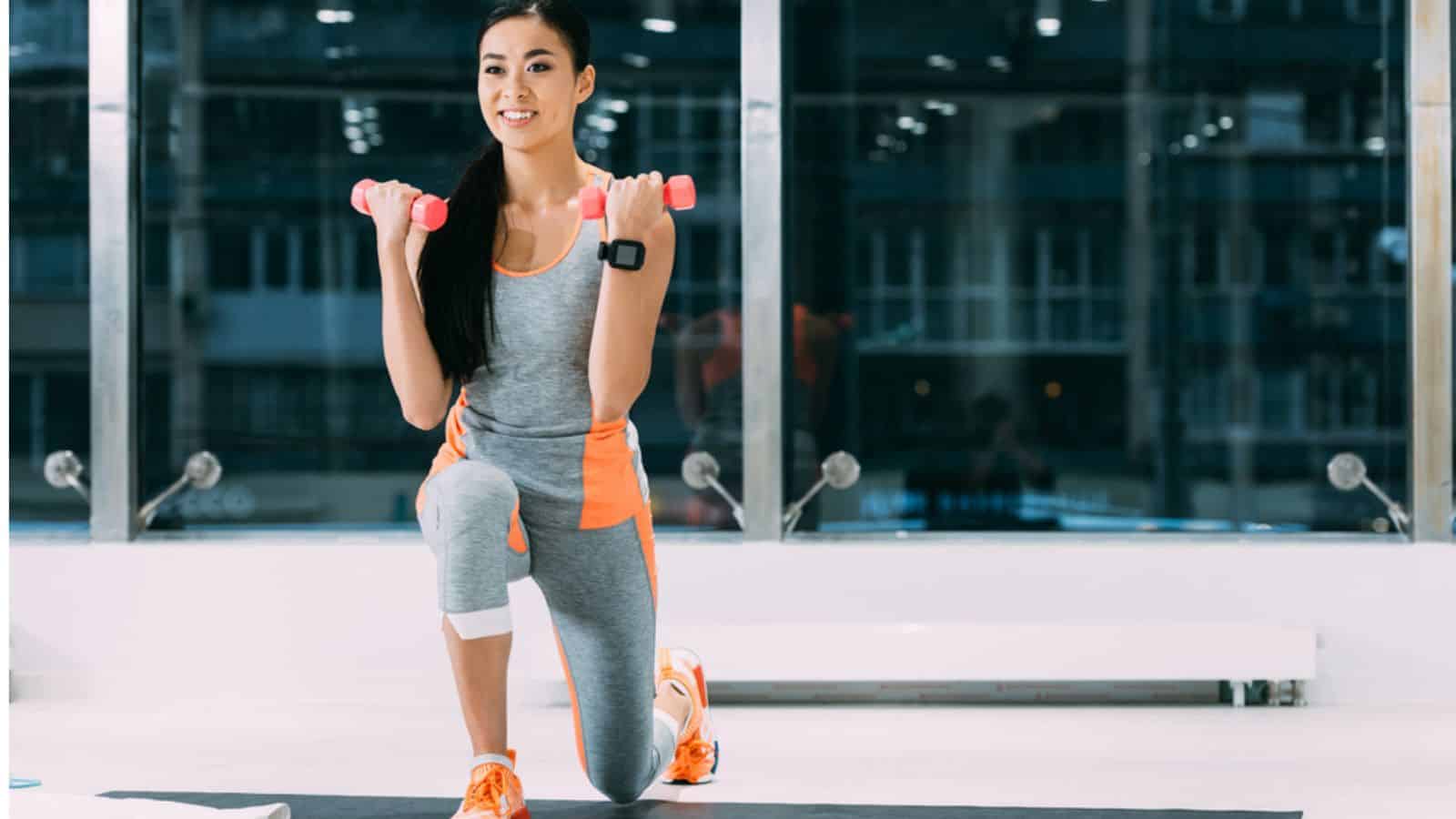 Smiling asian sportswoman doing exercise with dumbbells on fitness mat at gym