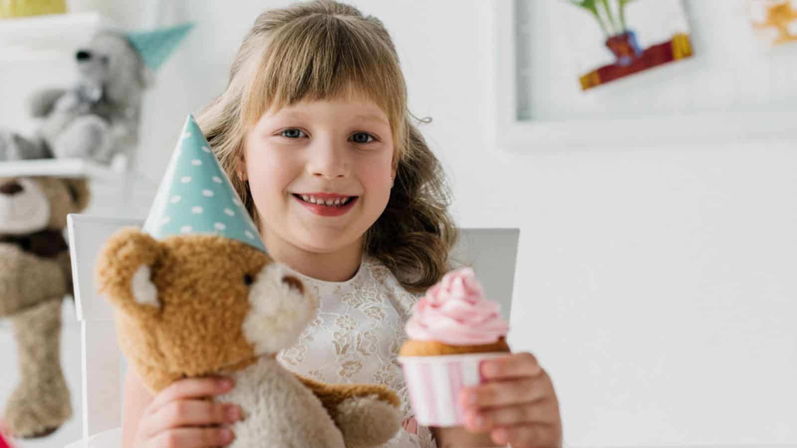 Smiling birthday kid showing cupcake and holding teddy bear
