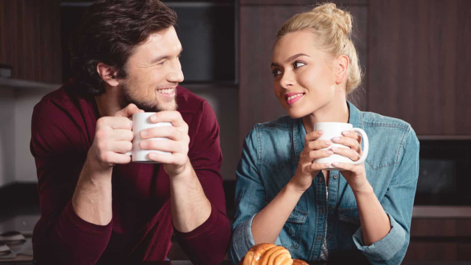 Smiling couple holding coffee cups and looking at each other in kitchen