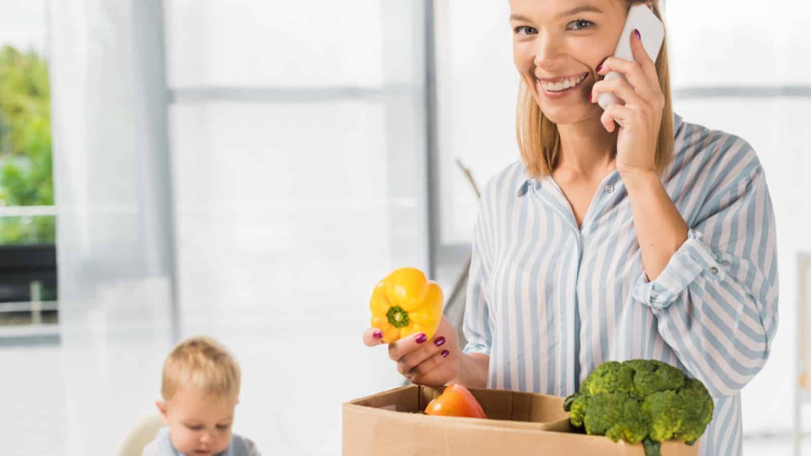 Smiling mother holding groceries while talking on smartphone