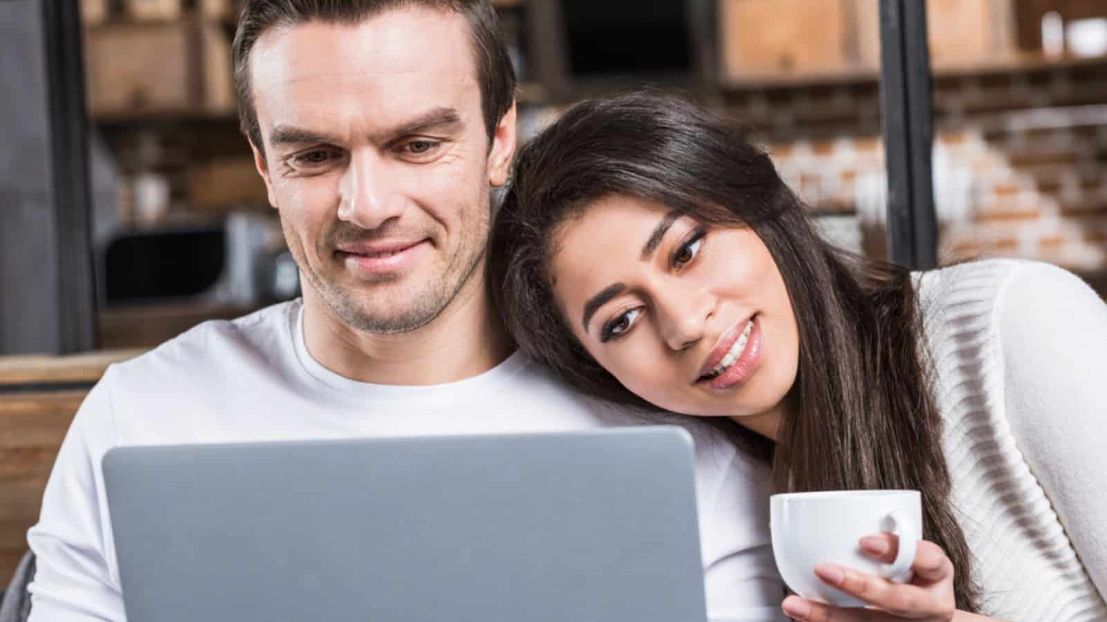 Smiling multiethnic couple using laptop together while woman drinking tea