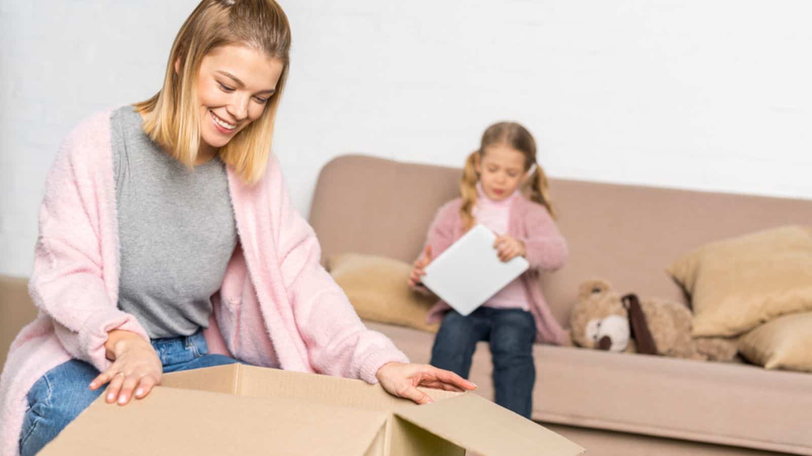 Smiling woman packing cardboard box while little daughter using digital