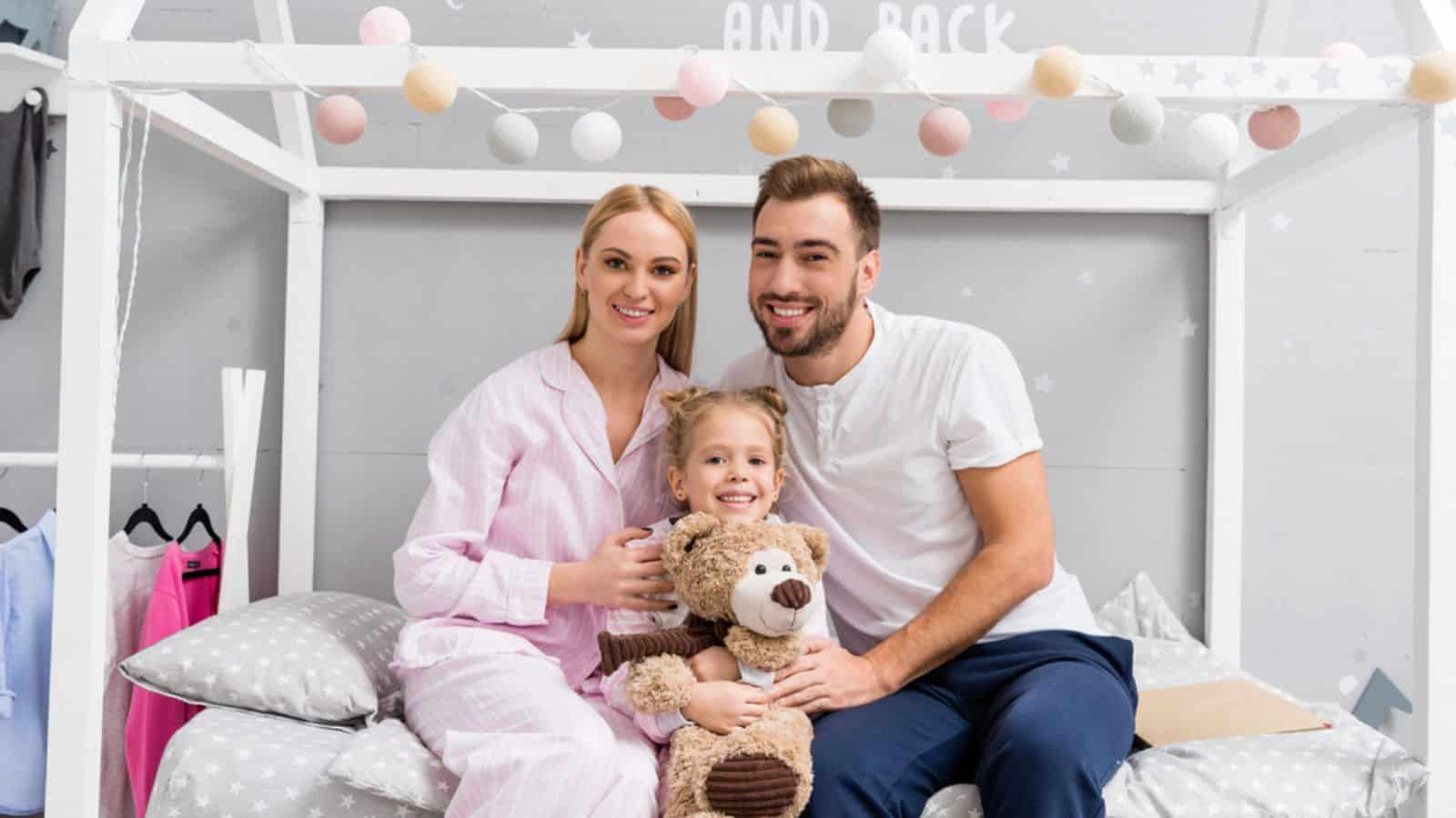 Smiling young family sitting on bed in kid bedroom and looking at camera