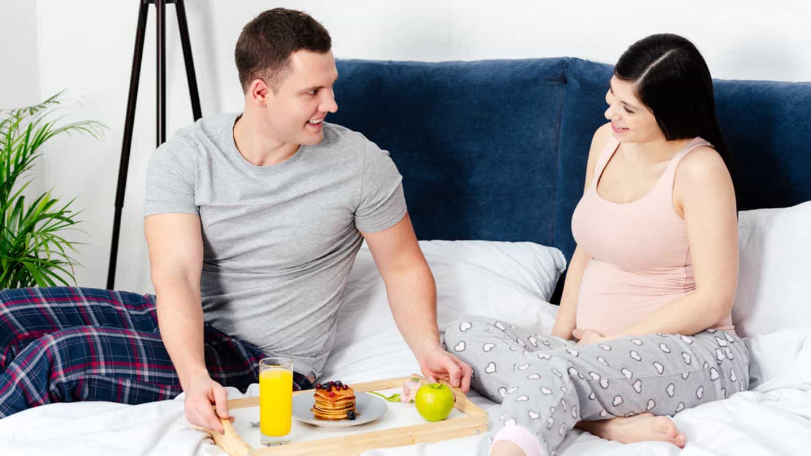 Smiling young man holding tray with breakfast and looking at happy pregnant women in bed