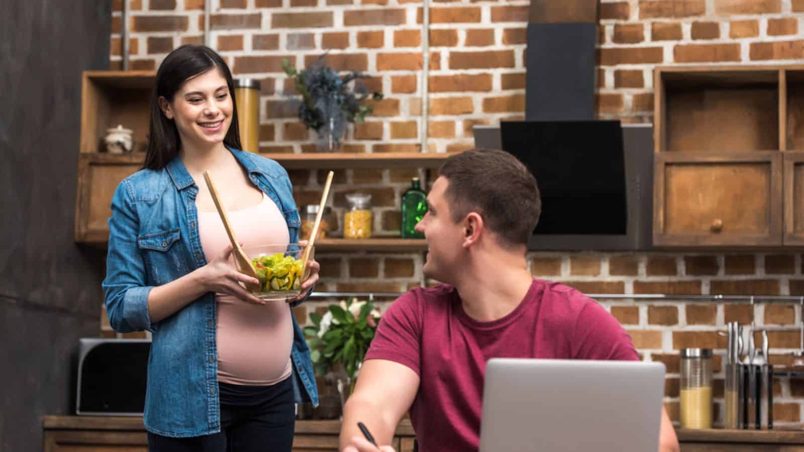 Smiling young man using laptop and looking at pregnant wife