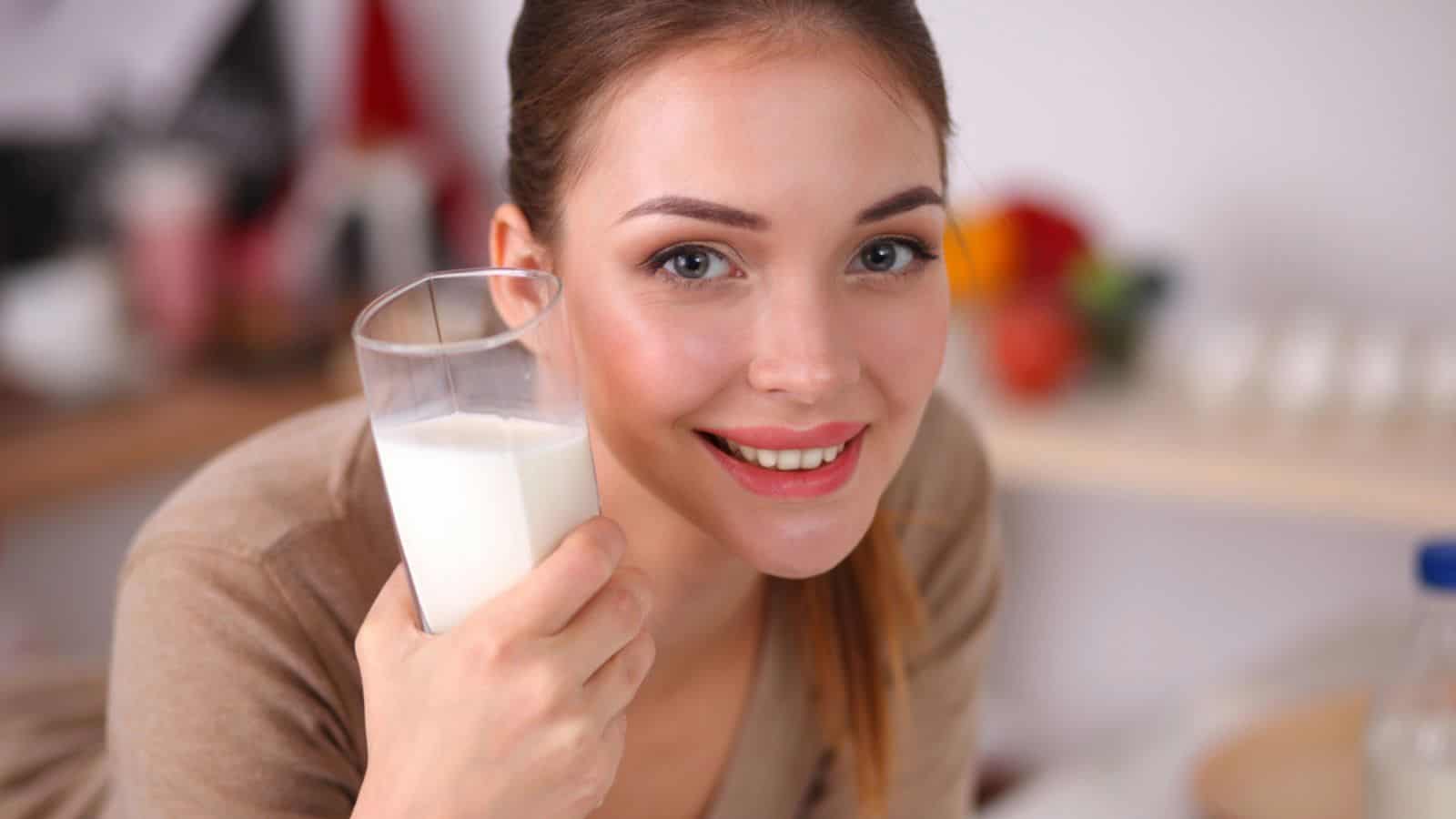 Smiling young woman drinking milk, standing in the kitchen