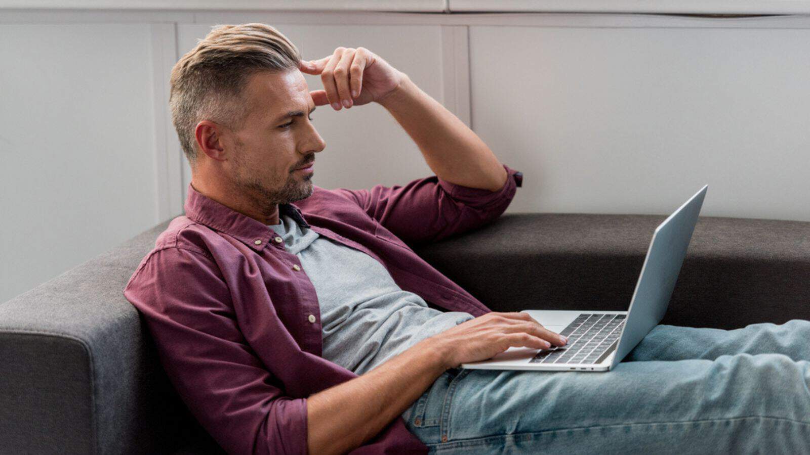 Thoughtful man laying on sofa and working on laptop at home office