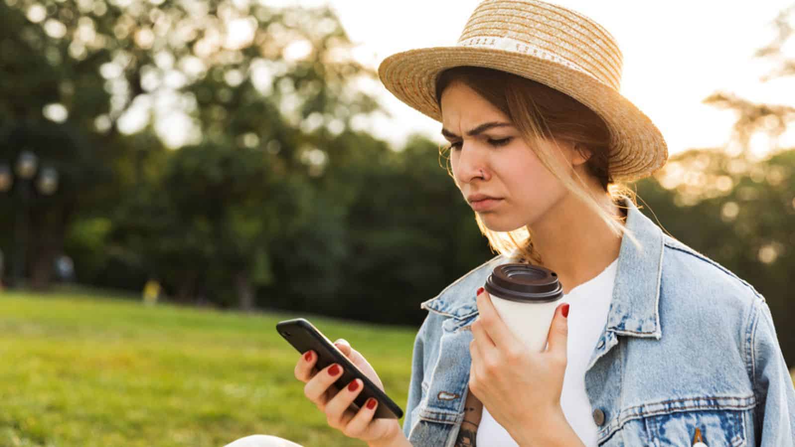 Upset young girl sitting on grass at the park, using mobile phone, holding cup oof coffee
