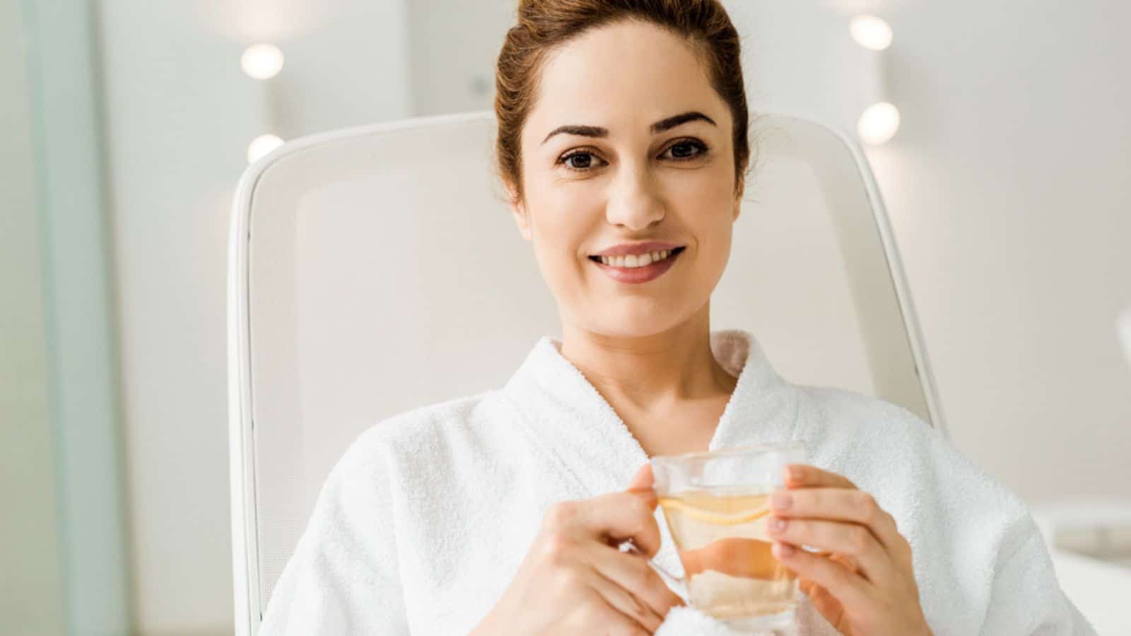 Woman in bathrobe holding cup with herbal drink and smiling at camera