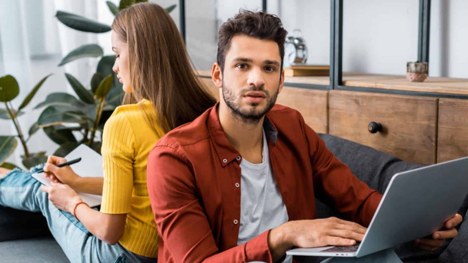 Young man holding laptop and sitting back to back with woman