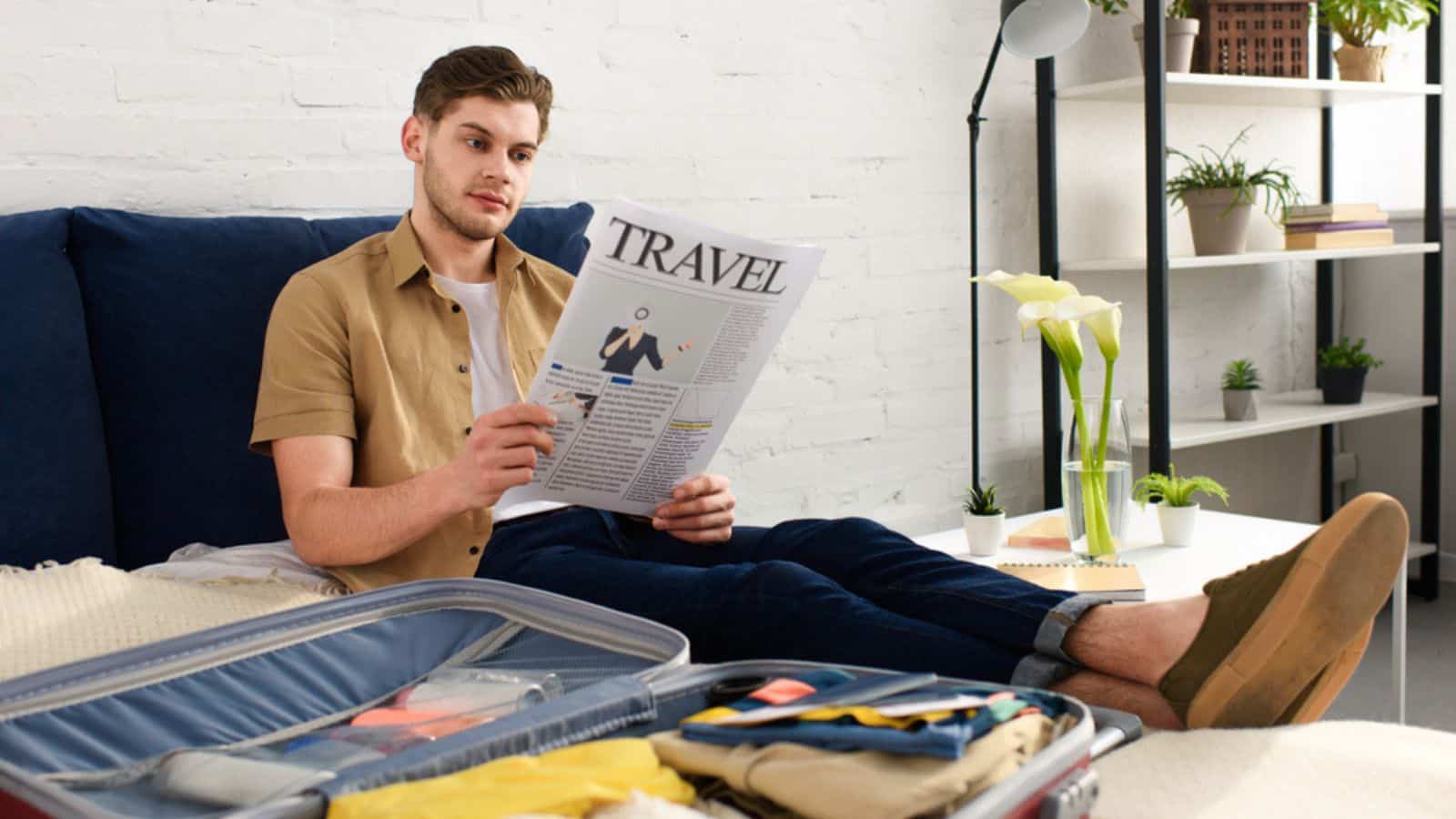 Young man reading travel newspaper while packing suitcase on bed